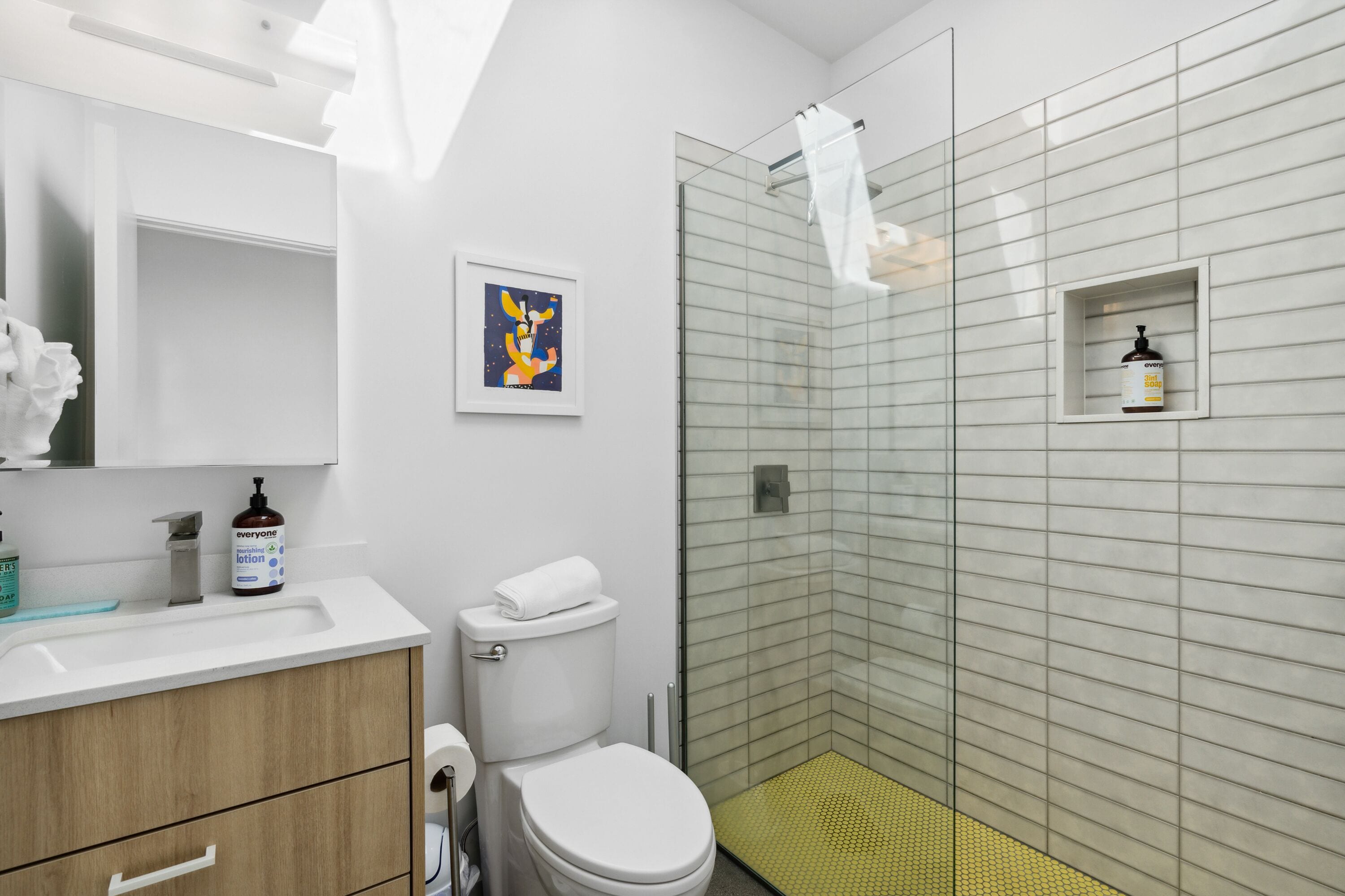 Une salle de bain moderne avec un lavabo, des toilettes et une douche, le tout de couleur blanche. Un tableau encadré est accroché au mur et un distributeur de savon est posé sur le lavabo.