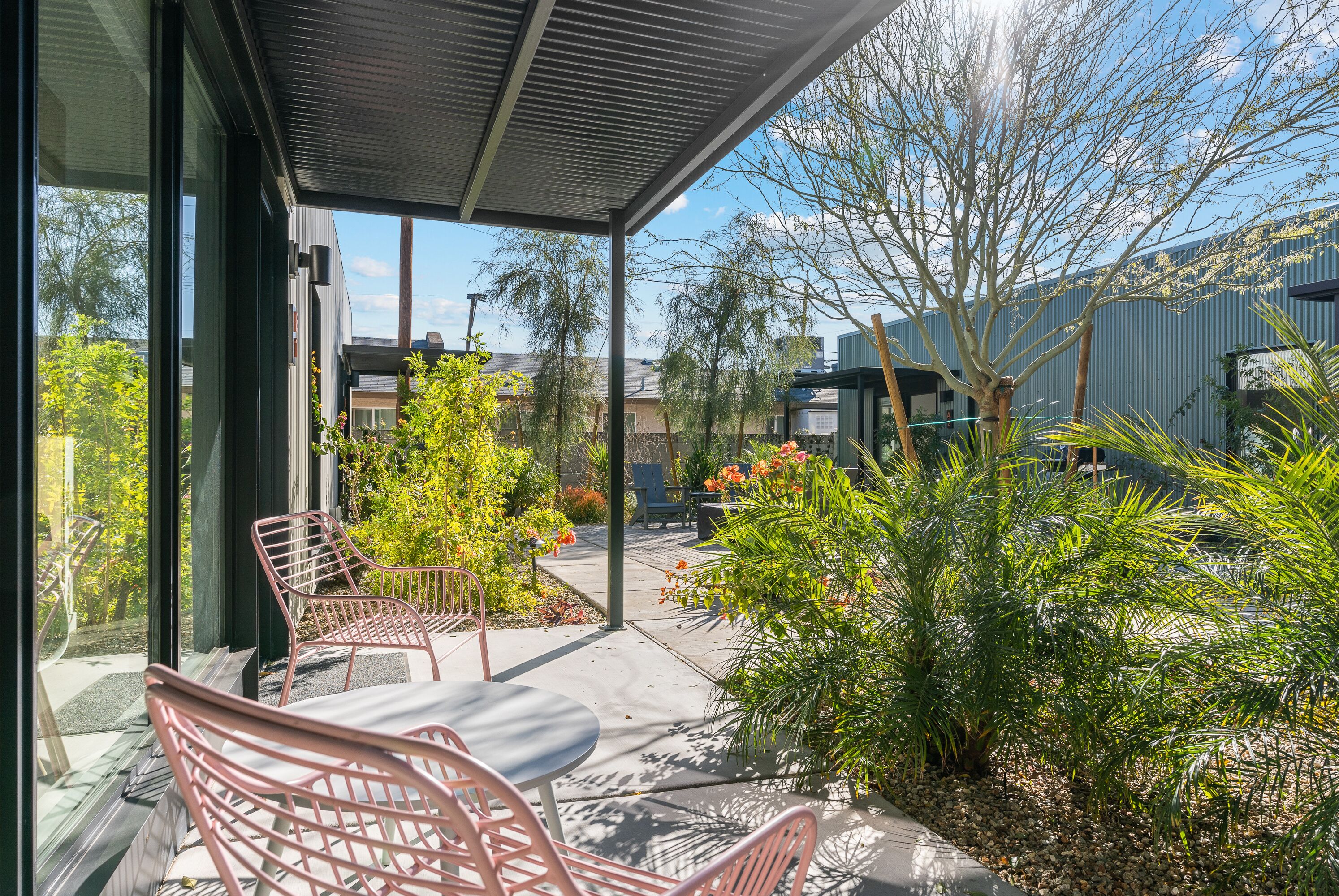 A covered outdoor seating area with metal chairs and a table, surrounded by greenery and a view of buildings in the background.