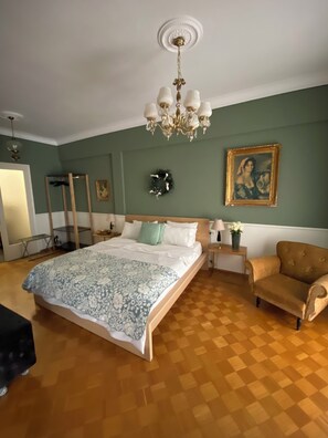 A bedroom with a large bed, a chandelier, a wooden floor, a chair, and a painting on the wall.
