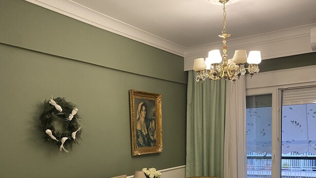 Un dormitorio con cama, mesita de noche, silla, candelabro, ventana con vistas y pared decorada con una corona y un cuadro enmarcado.