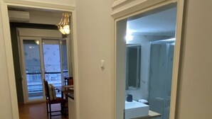 Un pasillo conduce a un baño con ducha, lavamanos y espejo. Hay una silla y una mesa en la habitación contigua.