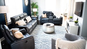 Uma sala de estar moderna com um sofá de couro preto, uma poltrona reclinável e uma mesa de centro. Há uma janela grande que oferece iluminação natural e uma TV de tela plana na parede.