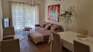A modern living room with a sofa, a dining table, and two chairs. There is a TV on the left, a chandelier, and a painting on the wall.