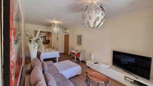 A modern living room with a large flat-screen TV, a sofa, a dining table with chairs, and a kitchen in the background.
