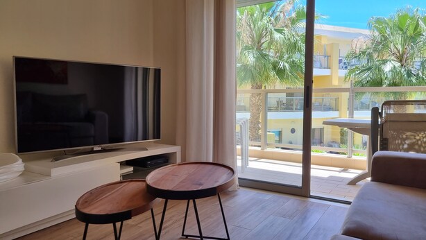 A modern living room with a flat-screen TV, two wooden side tables, and a large sliding glass door leading to a balcony with outdoor furniture.