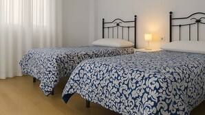 Una habitación con dos camas, cada una con una colcha estampada, una mesita de noche con lámpara y una ventana con cortinas traslúcidas.