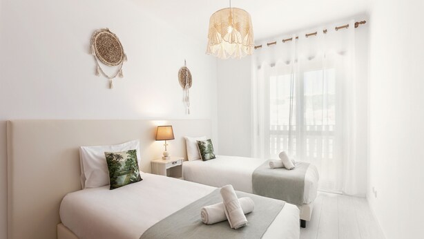 A hotel room with two beds, a bedside table with a lamp, a decorative wall hanging, and a large window with sheer curtains. - Escape 2 Nazaré - Private garage in the city (Nazaré)