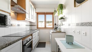 A modern kitchen with white cabinets, granite countertops, a built-in oven, and a window with a view of the city. - Escape 2 Nazaré - Private garage in the city (Nazaré)
