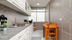 A compact kitchen with white cabinets, a granite countertop, a wooden dining table, and a window with frosted glass. - Cozy Retreat in the Southbank of Lisbon (Almada)
