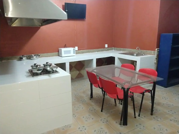 Une cuisine compacte équipée d’un four à micro-ondes, d’une cuisinière à gaz et d’un petit évier. On y trouve une table à manger en verre, entourée de chaises rouges.