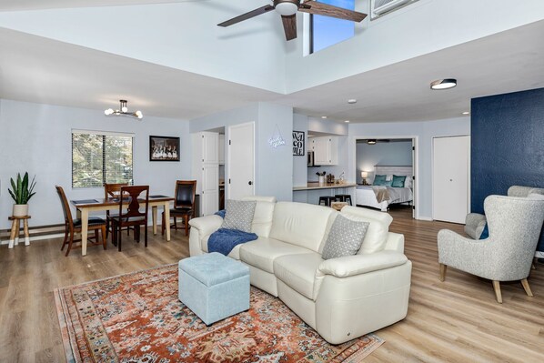 A spacious living area with a sofa, ottoman, and chairs, a dining table with chairs, and a kitchen in the background.