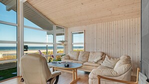 Ein geräumiges Zimmer mit großem Fenster mit Blick auf einen Strand, ein beiges Sofa, ein Holzstuhl und ein kleiner Tisch.