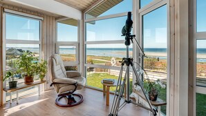 Ein geräumiges Zimmer mit großen Fenstern und Meerblick, ein bequemer Sessel, ein Teleskop und Topfpflanzen.