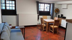 A compact room with a dining table, chairs, a radiator, a window with curtains, a fan, and a small kitchenette area.