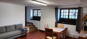 Un espace de vie compact comprenant un canapé, une petite table, une table à manger en bois avec des chaises, un radiateur et une fenêtre avec des rideaux.