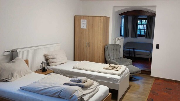 A small, well-lit room with two beds, a wardrobe, a chair, and a view of a dining area through an archway.