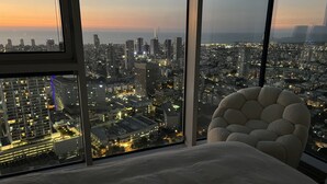 Une chambre d’hôtel située dans un immeuble de grande hauteur, dotée d’une grande fenêtre offrant une vue sur la ville au crépuscule.
