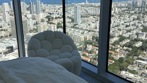 Une chambre située dans un immeuble de grande hauteur, dotée d’une grande fenêtre offrant une vue panoramique sur la ville, d’un fauteuil blanc moelleux et d’un lit parfaitement fait.