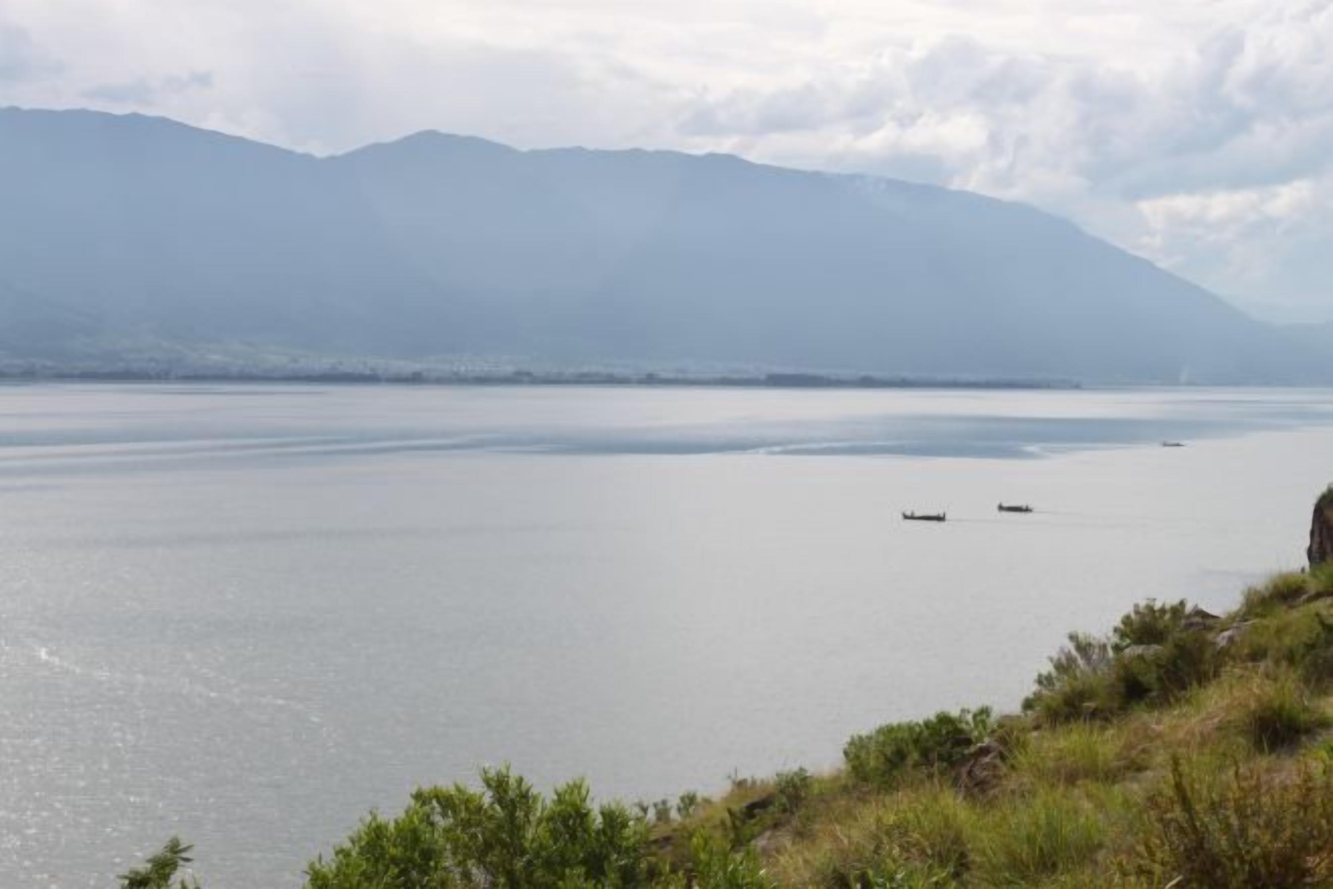 Água calma com barcos, uma cadeia montanhosa ao longe e uma margem gramada.