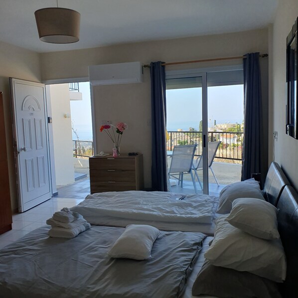 A bedroom with a bed, a dresser, a dresser with a vase of flowers, a balcony with a chair, and a view of the sea.