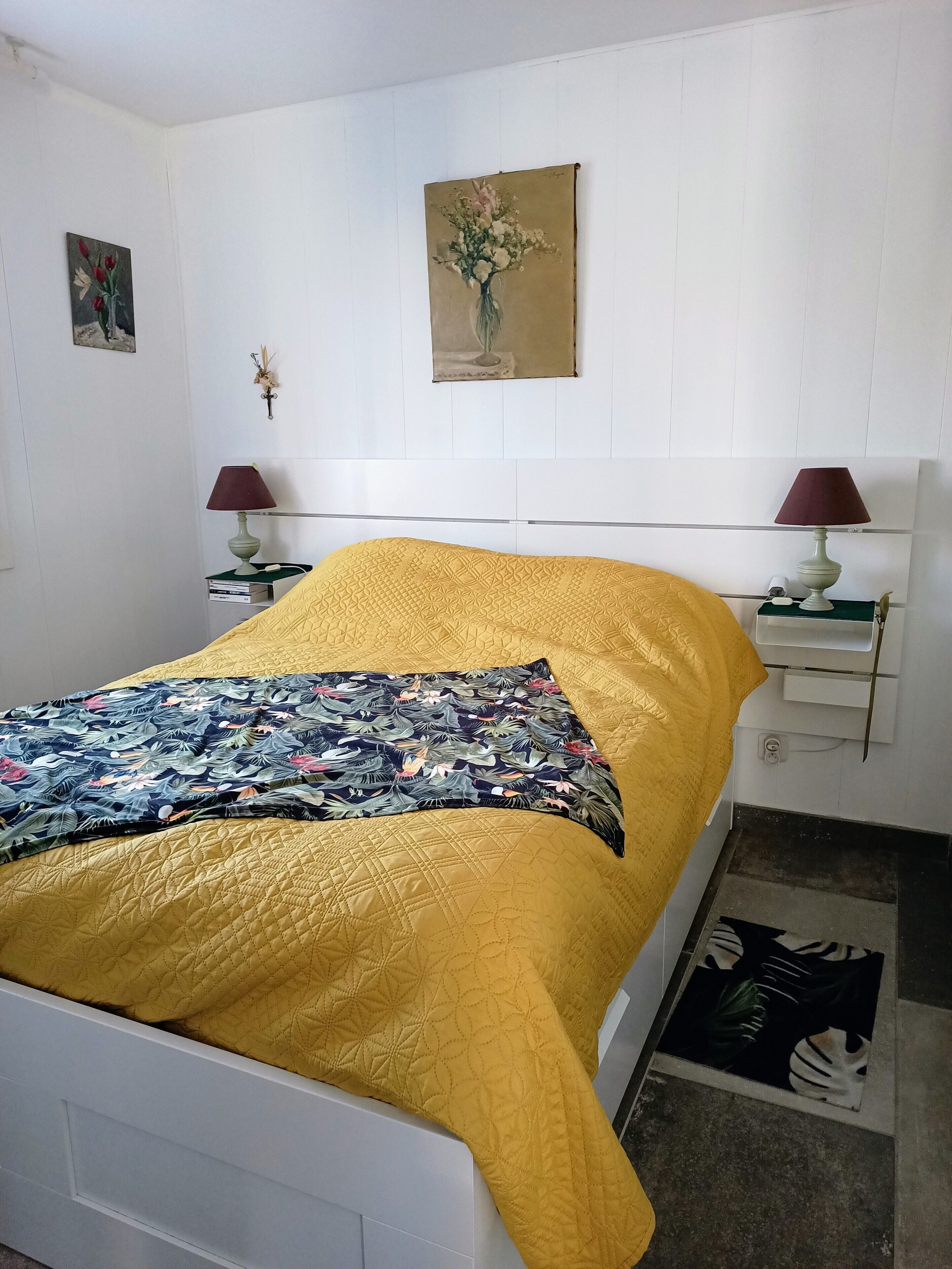 A bedroom with a bed, two bedside tables, a nightstand, a wall-mounted lamp, a painting, and a floral-patterned rug.