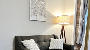A grey sofa with two white pillows, a floor lamp, a rug, and a wall art piece. - Tower Grove Studio - Fenced Yard, W/D, Pet-Friendly - 4 Mi to Downtown (St. Louis)