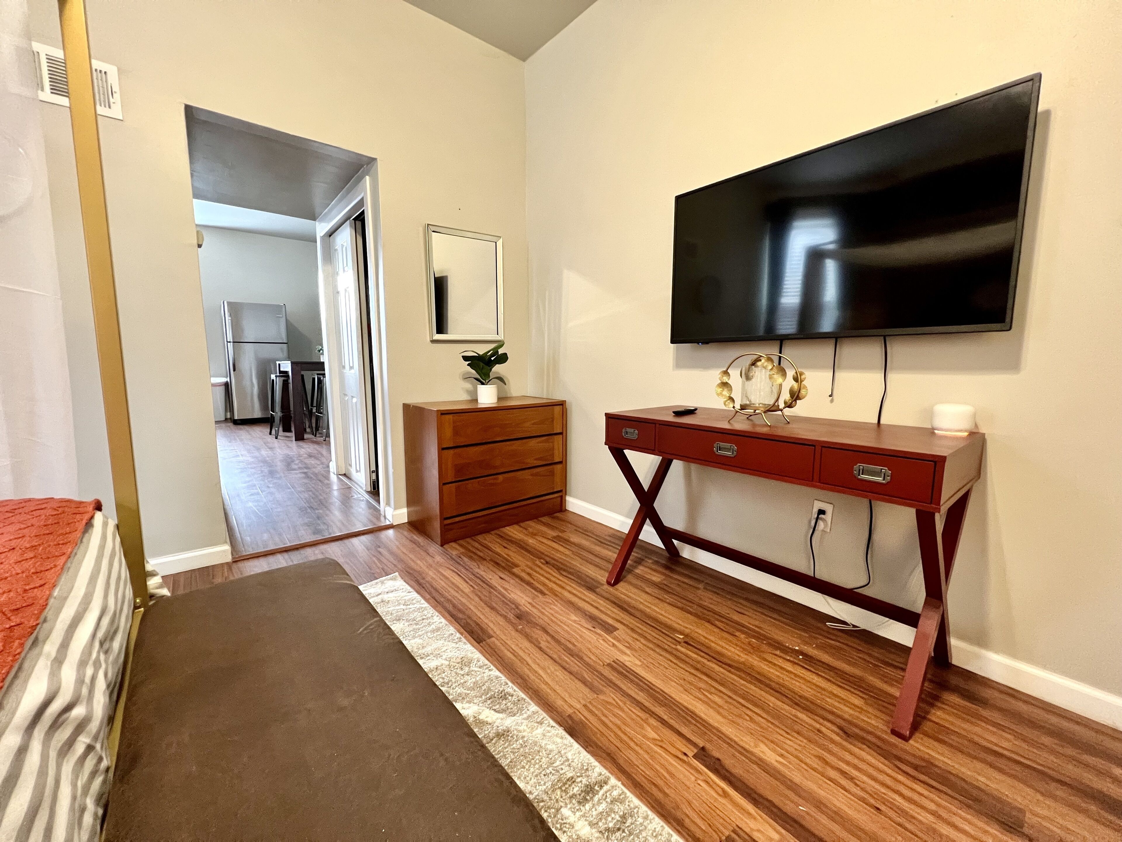 A room with a wooden floor, a red desk with drawers, a flat-screen TV mounted on the wall, and a small plant on the desk.