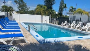 Una piscina con lettini blu e bianchi, un solarium piastrellato e una recinzione bianca sullo sfondo.