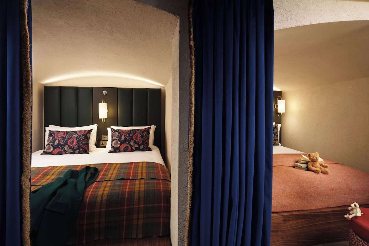 A hotel room with a bed, a nightstand, a teddy bear, and a red chair.