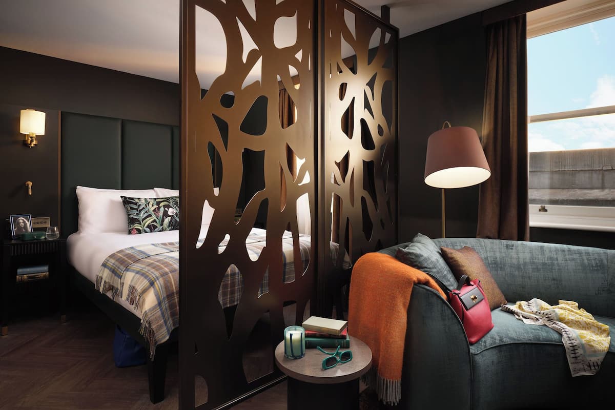 A hotel room with a bed, a sofa, a nightstand, a lamp, and a view of the sky.
