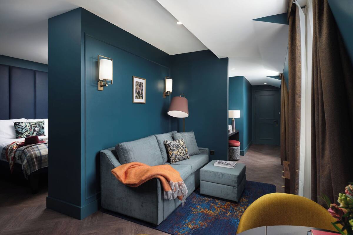 A modern hotel room with a grey sofa, a small ottoman, a red lamp, and a blue accent wall.