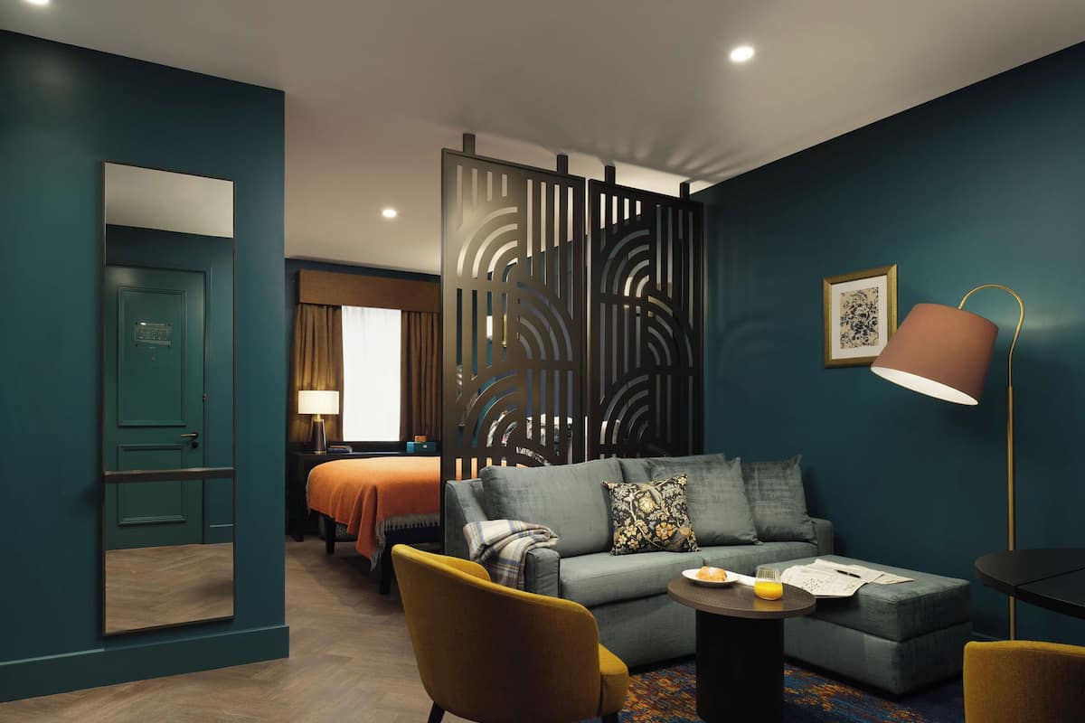 A modern hotel room with teal walls, a sofa set, a coffee table, and a lamp.