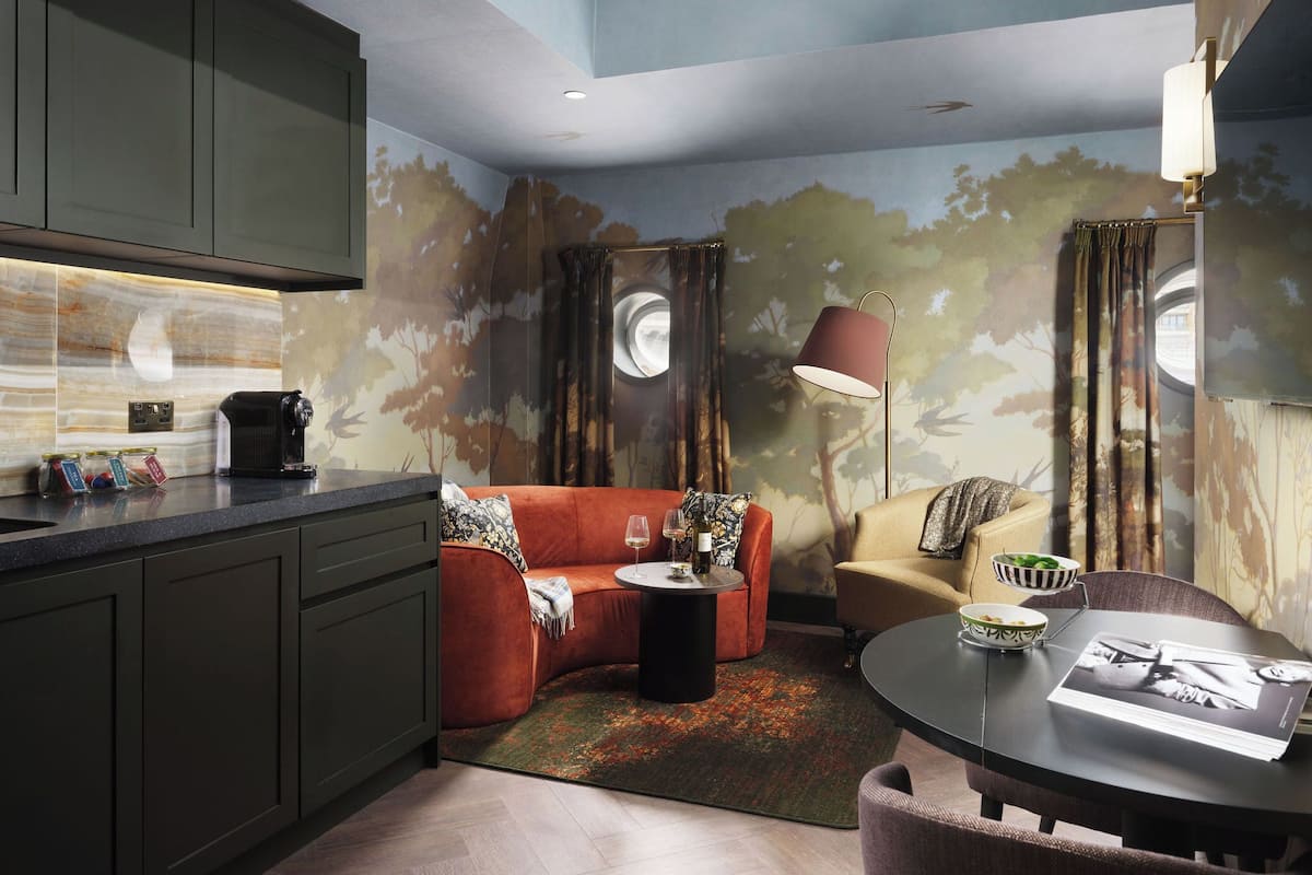 A modern living room with a sofa, a coffee table, and a dining table. There is a kitchen area with cabinets and a countertop. The room has a unique tree mural on the walls.