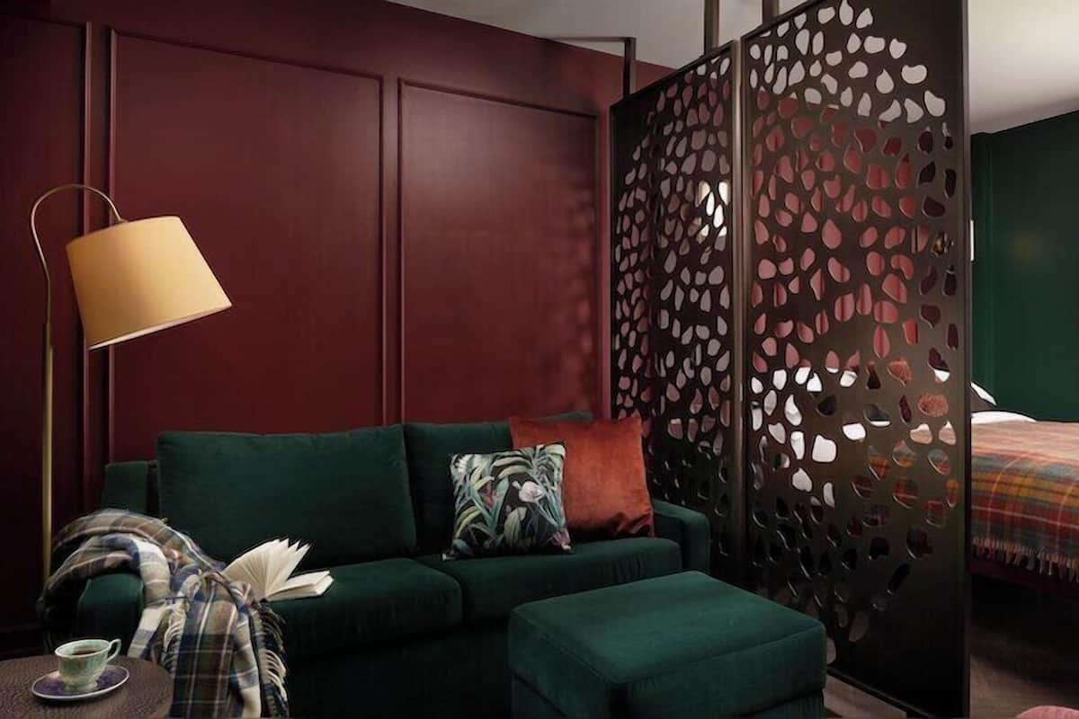 A hotel room with a green sofa, a patterned pillow, a plaid blanket, a side table with a cup, and a bed with a plaid blanket.