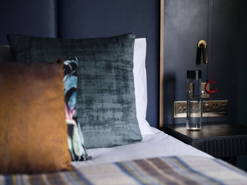 Una cama bien hecha con ropa de cama a rayas, un cabecero azul oscuro y una mesita de noche con un recipiente de vidrio y un objeto rojo.