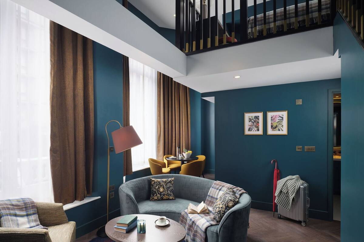 A modern hotel room with a blue accent wall, a sofa, a small table with books and a cup, a floor lamp, and a suitcase.