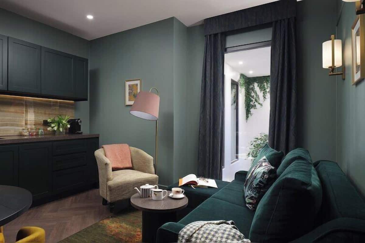 A modern living room with a dark green sofa, a round coffee table, a beige armchair, and a floor lamp. A kitchen with dark cabinets and a wooden countertop is visible in the background.