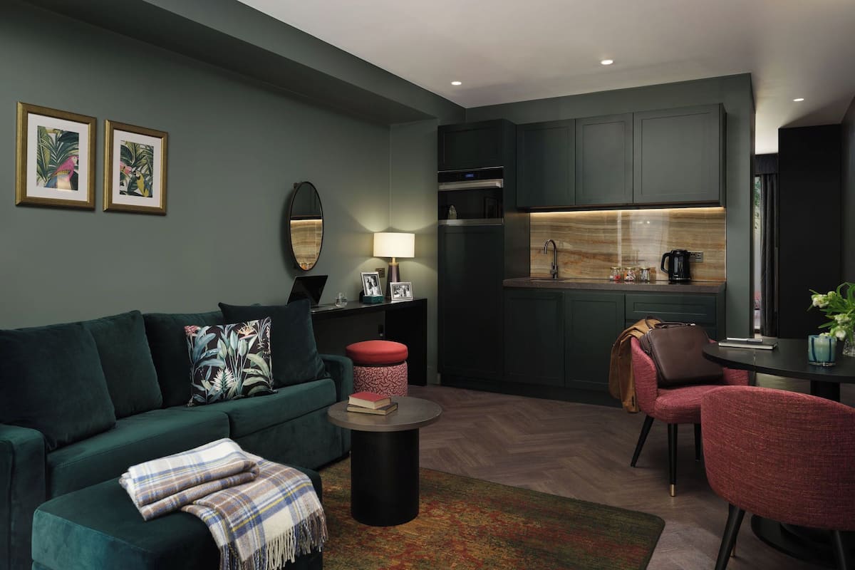 A modern living room with a dark green sofa, a red ottoman, and a coffee table. A kitchen with dark cabinets and a marble countertop is visible in the background.