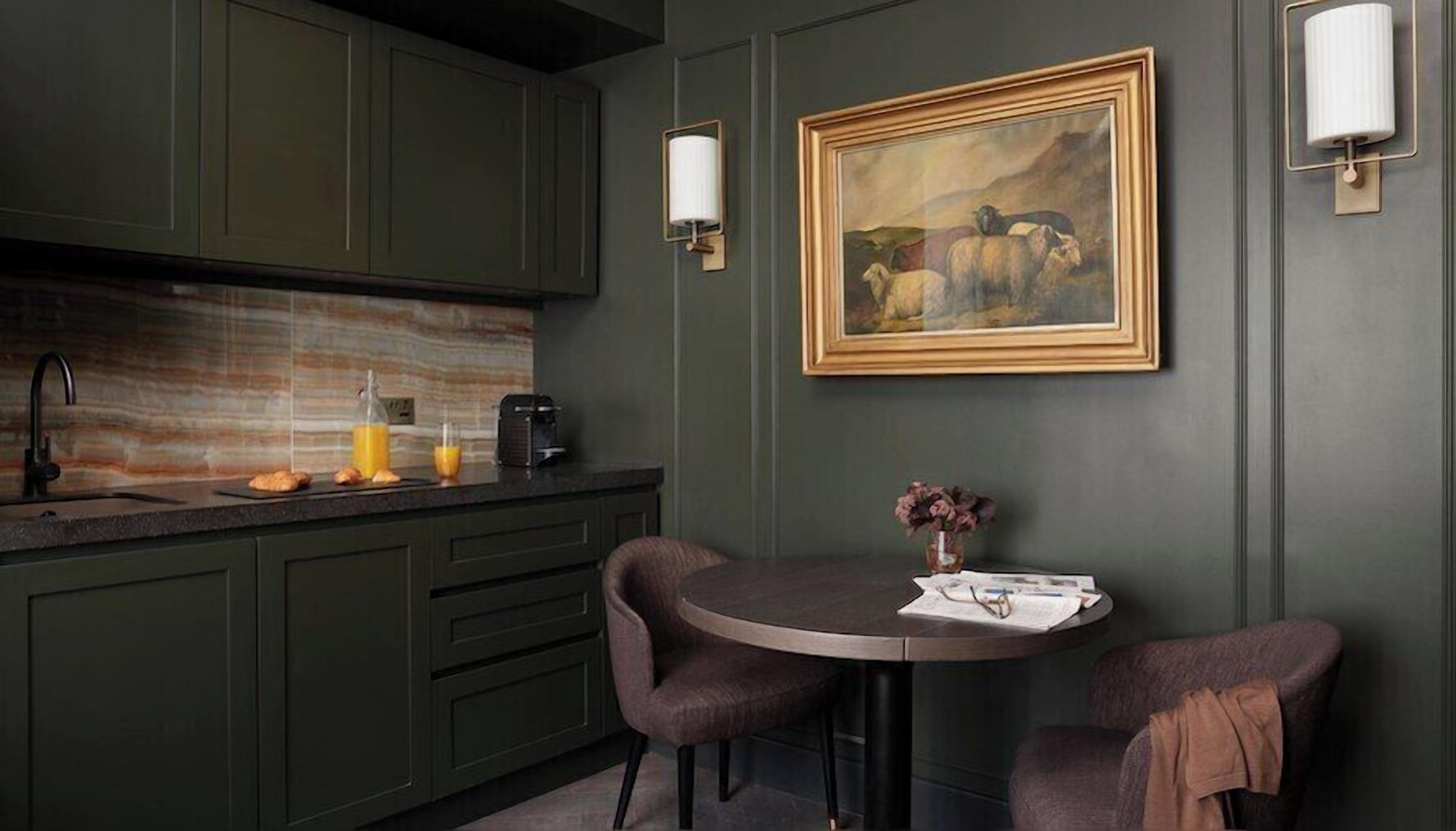 A modern kitchen with dark green cabinets, a striped backsplash, a round table with two chairs, and a framed painting on the wall.