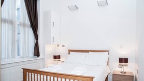 Une chambre à coucher avec un lit en bois, du linge de lit blanc, deux tables de chevet avec des lampes, une fenêtre avec des rideaux et un radiateur.