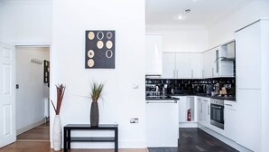 Une cuisine moderne avec des meubles blancs, des plans de travail noirs et des appareils en acier inoxydable. On aperçoit, au premier plan, un banc noir orné d’un vase et de plantes.
