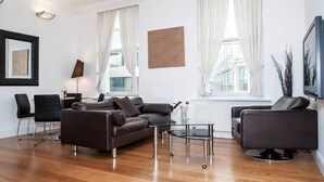 Un salon moderne avec un canapé en cuir marron, une table basse en verre et un fauteuil noir. On y trouve également deux chaises noires, une lampe et un tableau encadré accroché au mur.