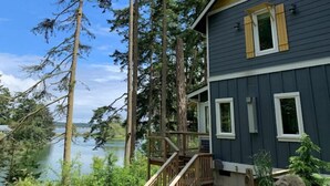 A two-story house with a deck overlooking a lake, surrounded by tall trees.