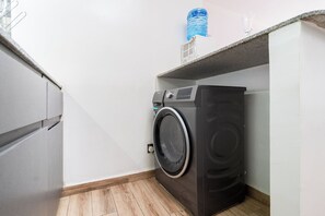 A compact laundry room with a washing machine, a small countertop, and a water dispenser. - Cozy 2-BR Aprt with Pool, Gym at Tatu City (Kiambu County)