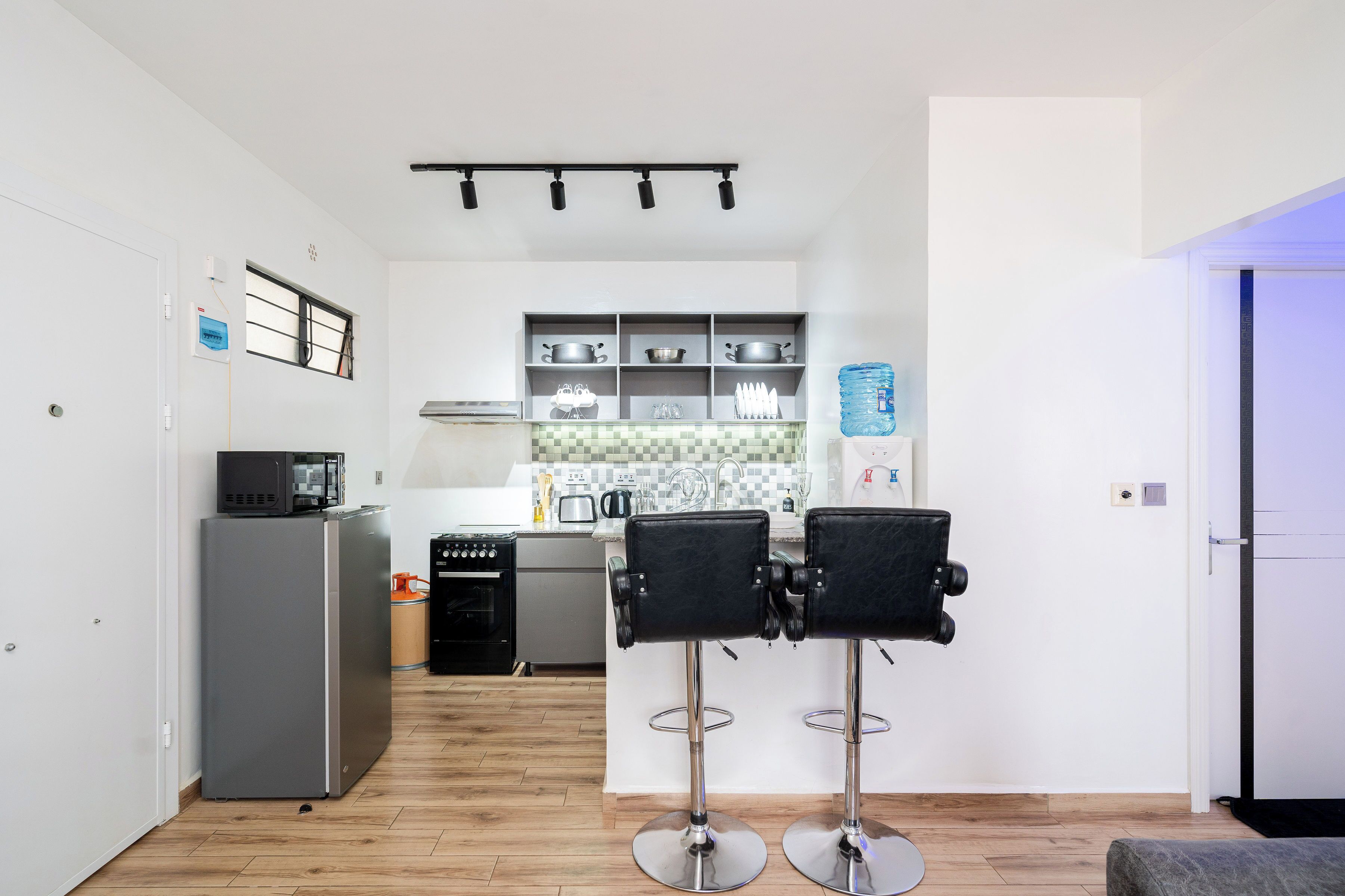A compact kitchen with a refrigerator, microwave, and coffee maker, two bar stools, and a tiled backsplash.