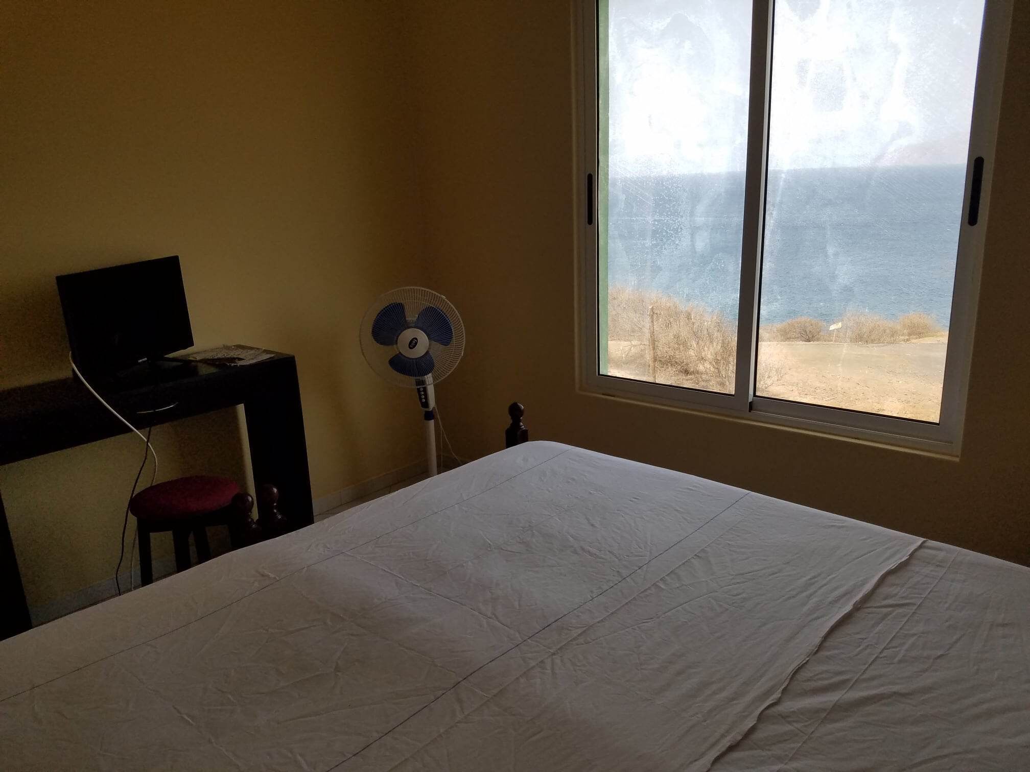 Una habitación con cama, escritorio con computadora, ventilador y ventana con vista a la playa.