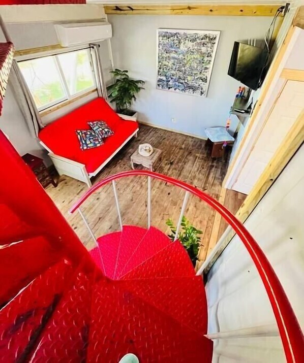 Una habitación con una escalera espiral roja, una cama con cubrecama rojo, una mesita, una planta y un televisor en la pared.