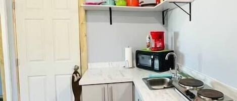 Eine kompakte Küche mit weißen Schränken, einer Mikrowelle, einer Kaffeemaschine und einem kleinen Regal mit bunten Gegenständen.