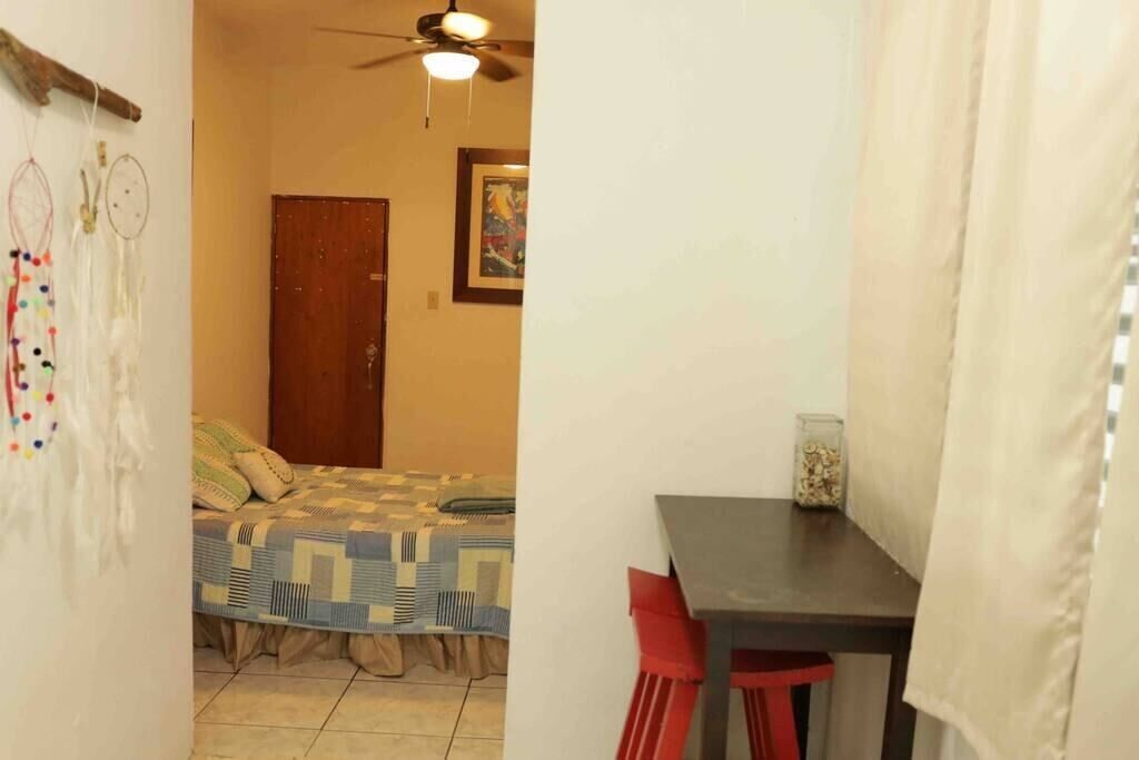 A small room with a bed, a desk, and a chair. There is a ceiling fan and a framed picture on the wall.