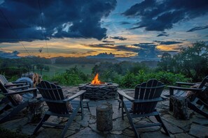 A stone fire pit with chairs around it, set against a backdrop of a sunset over a mountainous landscape.
